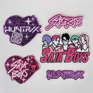 Könnte beinhalten: Eine Sammlung von aufgestickten Aufnähern mit den Schriftzügen "HUNTRAX" und "SAJA BOYS". Die Designs umfassen Herzformen, eine Gruppe von Cartoonfiguren und stilisierte Schriftzüge in Lila- und Rosatönen.