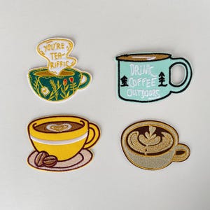 Op de afbeelding: Vier geborduurde patches met koffie- en theekopjesontwerpen. Eén patch zegt "You're Tea-riffic" boven een groene bloementheekop. Een andere zegt "Drink Coffee Outdoors" op een turquoise mok. Twee andere patches tonen koffiekopjes.
