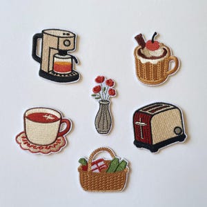 Op de afbeelding: Een verzameling van zes geborduurde patches met een beige koffiezetapparaat, een mok met slagroom en een kers, een vaas met rode bloemen, een kopje thee op een schoteltje, een rood met beige broodrooster en een bruine gevlochten picknickmand.