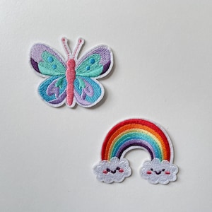 Op de afbeelding: Twee geborduurde patches: een vlinder en een regenboog. De vlinder heeft paarse, blauwgroene en blauwe vleugels met een roze lichaam. De regenboog patch heeft een kleurrijke boog met lachende wolken.
