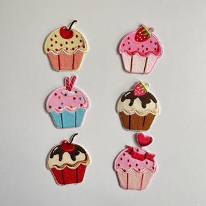 May include: A set of six embroidered cupcake patches featuring various designs including a cherry-topped yellow cupcake, a strawberry-topped pink cupcake, a blue cupcake with sprinkles, a chocolate-frosted cupcake, and a heart-topped pink cupcake.