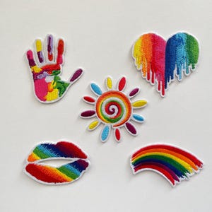 Könnte beinhalten: Eine Sammlung gestickter Aufnäher mit farbenfrohen Designs. Enthält einen Handabdruck, ein Herz mit tropfenden Regenbogenfarben, eine Sonne mit einem spiralförmigen Zentrum, ein Lippenpaar und einen Regenbogenbogen. Jeder Aufnäher hat einen weißen Rand.