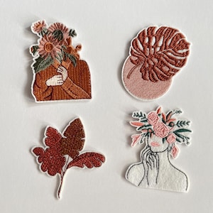 Può includere: Quattro toppe ricamate con disegni floreali e botanici. Una toppa presenta una figura con fiori al posto della testa, un'altra una foglia di monstera, e altre due presentano composizioni floreali e di foglie. Le toppe sono nei toni del marrone, rosa e verde.