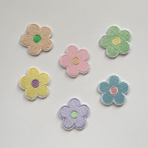Könnte beinhalten: Sechs gestickte Blumenaufnäher in Pastellfarben: Beige, Rosa, Grün, Gelb, Blau und Lavendel. Jede Blume hat eine kontrastierende Mitte und einen weißen Rand. Diese Aufnäher eignen sich zum Verzieren von Kleidung oder Accessoires.