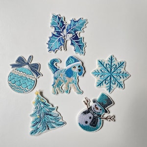 Könnte beinhalten: Eine Sammlung von gestickten Aufnähern mit winterlichem Feiertags-Thema. Die Aufnäher zeigen eine blau-weiße Schneeflocke, einen Weihnachtsbaum, einen Schneemann, einen Pudel mit Hut, ein Stechpalmenblatt und eine Weihnachtskugel.