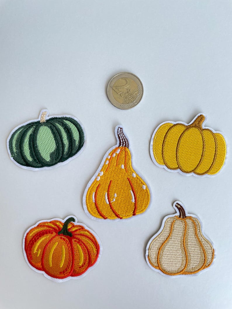 May include: Five embroidered pumpkin patches in various shapes and colors, including green, yellow, orange, and cream. Each patch has a white border and detailed stitching. A two-euro coin is also visible.