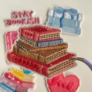 May include: Embroidered patches depicting stacks of books in shades of red, brown, and blue. Other patches include a heart with the word "Love" and the phrase "Stay Bookish". The patches are set against a white backdrop.