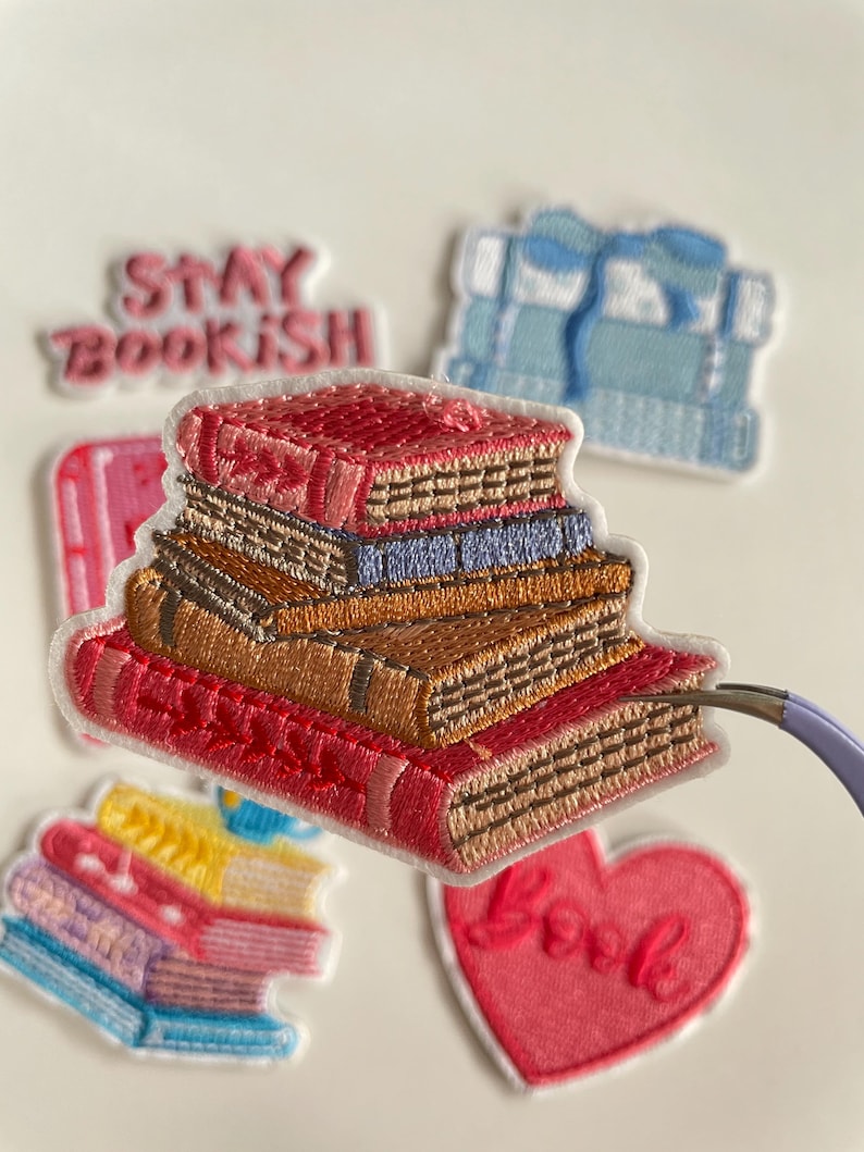 May include: Embroidered patches depicting stacks of books in shades of red, brown, and blue. Other patches include a heart with the word "Love" and the phrase "Stay Bookish". The patches are set against a white backdrop.