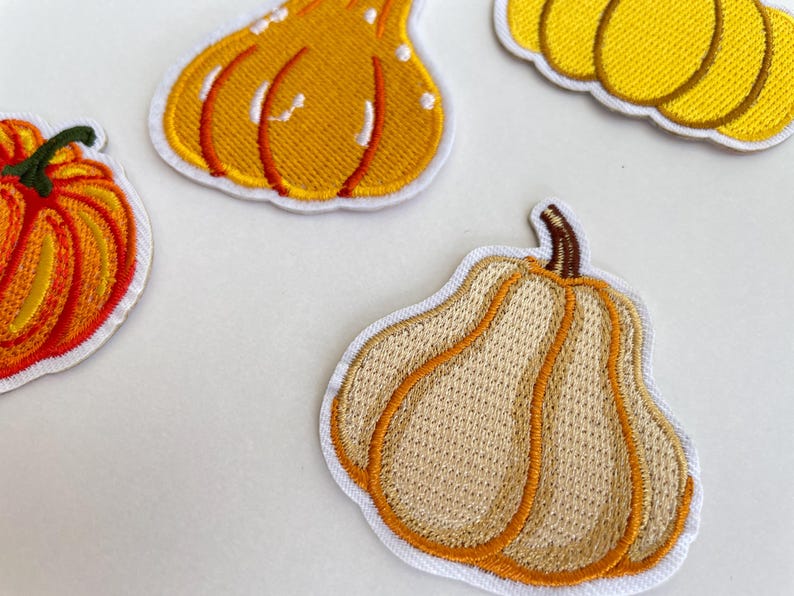 May include: Embroidered patches featuring various pumpkin designs in shades of orange, yellow, and tan. Each patch has a white border and detailed stitching, perfect for fall-themed crafts and decorations.