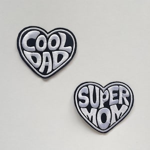 Könnte beinhalten: Zwei herzförmige Aufnäher. Einer zeigt in weißen Buchstaben "COOL DAD" auf schwarzem Grund. Der andere zeigt in weißen Buchstaben "SUPER MOM" auf schwarzem Grund.