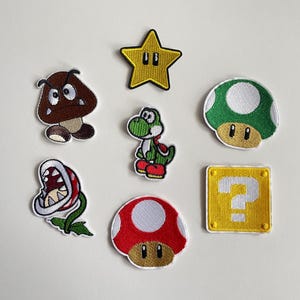 以下が含まれることがあります： スーパーマリオのビデオゲームシリーズのキャラクターとシンボルをあしらった刺繍パッチのコレクション。黄色い星、緑のキノコ、赤いキノコ、クエスチョンマークブロック、クッパ、ヨッシー、パックンフラワーが含まれています。