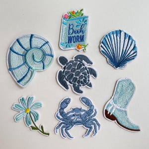Peut inclure: Écussons brodés sur le thème de la mer, dont un coquillage, un livre avec le texte "Book Worm", une coquille, une tortue, une fleur, un crabe et une botte. Les écussons sont bleus, blancs et marron.