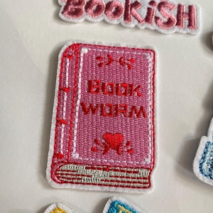 May include: A pink embroidered patch in the shape of a book, featuring the words "BOOK WORM" in white. The book has red stitching and a red heart design. The patch is set against a white backdrop.