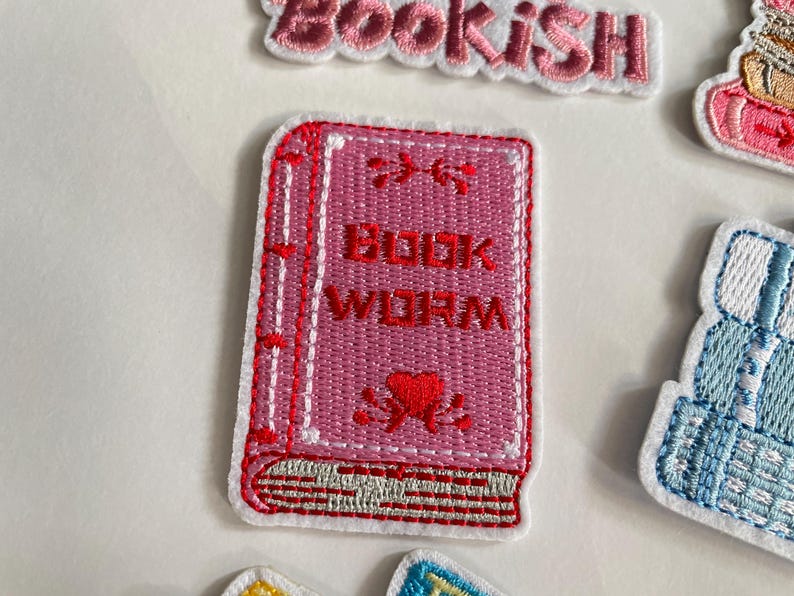 May include: A pink embroidered patch in the shape of a book, featuring the words "BOOK WORM" in white. The book has red stitching and a red heart design. The patch is set against a white backdrop.