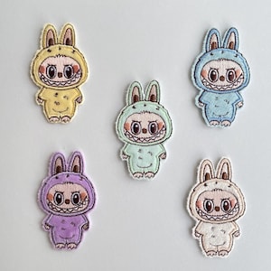 Könnte beinhalten: Fünf gestickte Aufnäher mit einer Cartoonfigur in einem Hasenkostüm. Die Aufnäher sind in Gelb, Blau, Grün, Lila und Weiß gehalten. Jeder Aufnäher hat ein lächelndes Gesicht mit großen Augen und einem grinsenden Mund. Die Aufnäher befinden sich auf weißem Hintergrund.