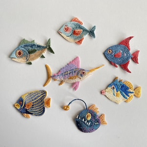 Könnte beinhalten: Eine Sammlung von sieben gestickten Fischaufnähern, die verschiedene Arten darstellen, darunter einen Schwertfisch, einen Anglerfisch und mehrere tropische Fische in leuchtenden Farben wie Blau, Gelb, Rosa und Orange, alle mit detaillierter Stickerei.