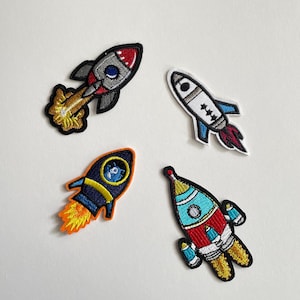 Può includere: Quattro toppe ricamate a forma di razzo in vari colori. Un razzo è rosso, grigio e nero con una fiamma gialla. Un altro è bianco, blu e rosso. Il terzo è blu navy, arancione e giallo. L'ultimo è rosso, blu e giallo.