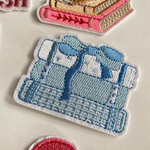 May include: Embroidered patch showing a stack of light blue books tied with a blue ribbon. The patch has a white border and detailed stitching, suggesting a book-themed design. Other patches are visible.
