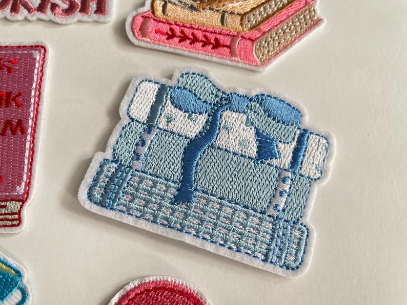 May include: Embroidered patch showing a stack of light blue books tied with a blue ribbon. The patch has a white border and detailed stitching, suggesting a book-themed design. Other patches are visible.