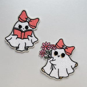 May include: Two embroidered patches of ghost characters. One reads a book, the other holds flowers. Both have a red bow with a skull detail. The patches are white with black and red accents. Ideal for clothing or accessories.