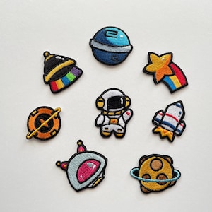 Könnte beinhalten: Eine Sammlung von gestickten Weltraum-Patches. Designs umfassen einen blauen Planeten, eine Rakete, einen Sternschnuppen, einen Astronauten, eine Raumstation und einen Planeten mit einem Ring. Die Patches sind farbenfroh und detailliert.