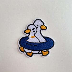 May include: Embroidered patch featuring two cartoon ducks. One duck is inside a blue inflatable ring, and the other duck is behind it. The ducks have white bodies, yellow beaks and feet, and black eyes. The patch has a white border.