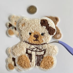 Pode incluir: Close-up de um patch de urso de pel&uacute;cia com corpo creme e detalhes marrons, incluindo um la&ccedil;o. O urso tem uma textura fofa. Outros patches semelhantes est&atilde;o no fundo. Uma moeda e uma pin&ccedil;a tamb&eacute;m s&atilde;o vis&iacute;veis.