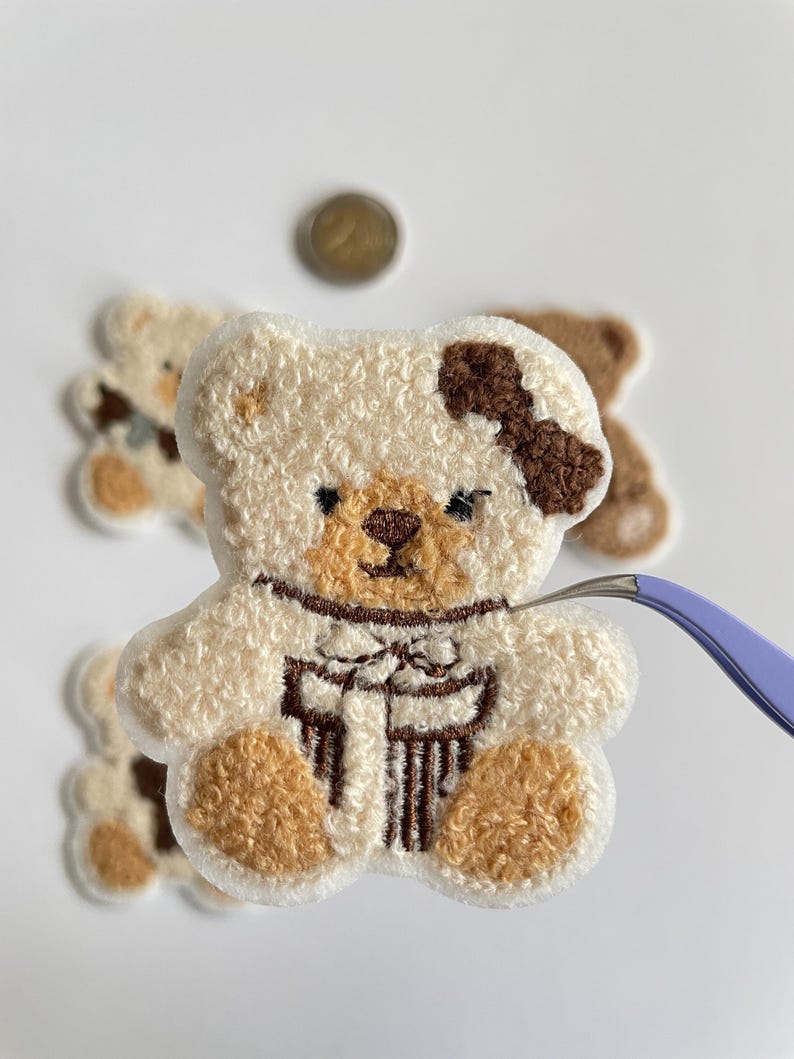 Pode incluir: Close-up de um patch de urso de pel&uacute;cia com corpo creme e detalhes marrons, incluindo um la&ccedil;o. O urso tem uma textura fofa. Outros patches semelhantes est&atilde;o no fundo. Uma moeda e uma pin&ccedil;a tamb&eacute;m s&atilde;o vis&iacute;veis.