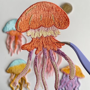 May include: Embroidered jellyfish patches in various colors, including orange, pink, purple, yellow, and blue. The patches feature detailed stitching and are held with tweezers. The jellyfish have a dome-shaped top and flowing tentacles, perfect for crafting projects.