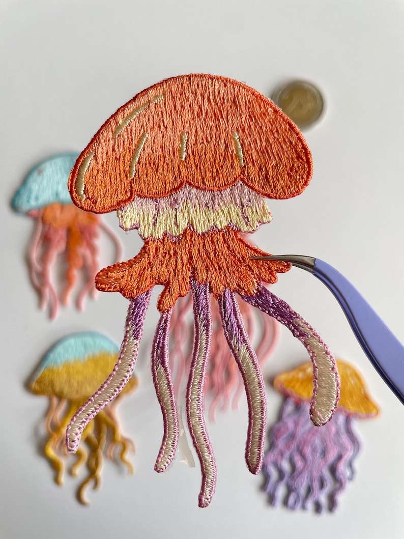 May include: Embroidered jellyfish patches in various colors, including orange, pink, purple, yellow, and blue. The patches feature detailed stitching and are held with tweezers. The jellyfish have a dome-shaped top and flowing tentacles, perfect for crafting projects.