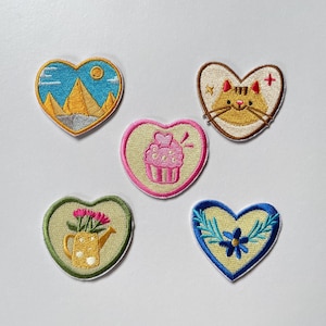Könnte beinhalten: Fünf herzförmige Stickaufnäher. Einer zeigt gelbe Pyramiden unter blauem Himmel, einer ein beiges Katzengesicht, einer einen rosa Cupcake, einer eine gelbe Gießkanne mit rosa Blumen und einer eine blaue Blume mit grünen Blättern auf cremefarbenem Grund.