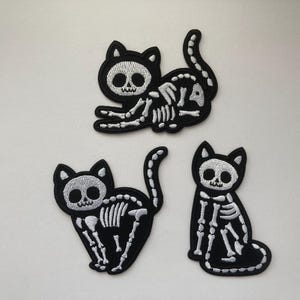 Peut inclure: Trois écussons noirs en forme de chat avec des motifs de squelettes blancs. Les chats sont dans différentes poses : un couché, un assis et un avec la queue en l'air. Idéal pour les projets sur le thème d'Halloween ou gothique.