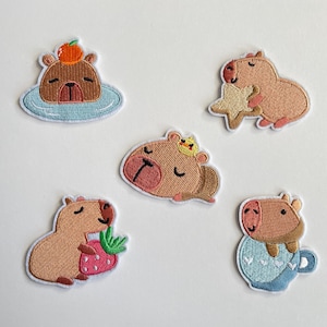 May include: Set of five embroidered patches featuring cartoon capybaras in various poses. One capybara is in a blue pool with an orange on its head. Another holds a star, and another eats a strawberry. The others are in a crown and a teacup.