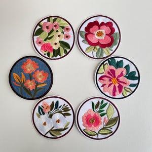 Könnte beinhalten: Eine Sammlung von sieben gestickten Blumenaufnähern. Jeder runde Aufnäher zeigt ein anderes Blumendesign in verschiedenen Farben, darunter Rosa, Orange und Weiß, mit grünen Blättern. Die Aufnäher haben einen weißen Hintergrund und einen dunklen Rand.