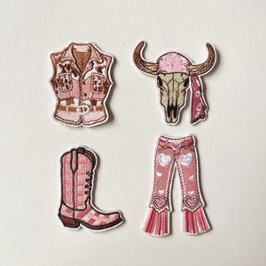 Puede incluir: Un conjunto de cuatro parches bordados que incluye un chaleco rosa con detalles marrones, un cráneo de toro beige con un pañuelo rosa, una bota vaquera a cuadros rosas y blancos, y pantalones rosas acampanados con corazones blancos.