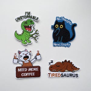 Può includere: Quattro toppe ricamate con disegni umoristici. Una presenta un dinosauro verde con la scritta "I'M UNSTOPPABLE". Un'altra mostra un gatto nero con la scritta "No Thoughts Head Empty". Un gatto bianco con "NEED MORE COFFEE" e un dinosauro marrone addormentato con "TIREDSAURUS" completano il set.