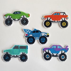 Könnte beinhalten: Fünf gestickte Monster-Truck-Patches in verschiedenen Farben: Grün, Orange, Blau, Türkis und Lila. Jeder Patch hat große schwarze Räder mit weißen Akzenten und einem weißen Rand. Die Trucks haben unterschiedliche Designs.
