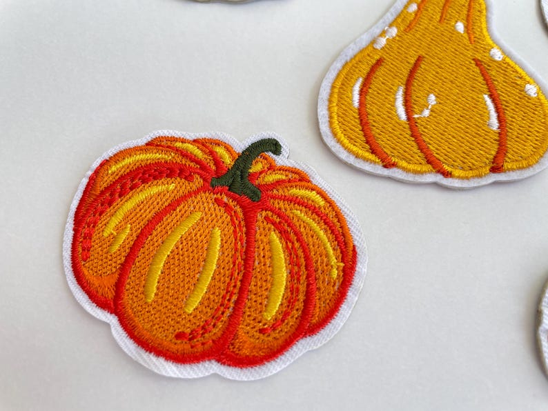 May include: Embroidered patches featuring a pumpkin and a gourd. The pumpkin patch is orange and red with a green stem. The gourd patch is yellow with white accents. Both patches have a white border.