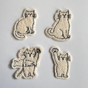 Könnte beinhalten: Vier cremefarbene Katzenaufnäher mit schwarzen gestickten Konturen. Zwei Katzen sitzen, eine winkt und eine andere umarmt einen Fisch. Diese Aufnäher sind ideal, um Kleidung oder Accessoires einen Hauch von Katzencharme zu verleihen.