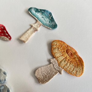 May include: Embroidered mushroom patches in various colors, including red, blue, and orange. The patches feature detailed stitching and are likely for decorative purposes, such as adding to clothing or accessories. The patches are on a white background.
