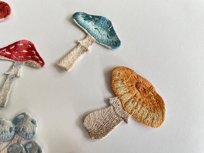 May include: Embroidered mushroom patches in various colors, including red, blue, and orange. The patches feature detailed stitching and are likely for decorative purposes, such as adding to clothing or accessories. The patches are on a white background.