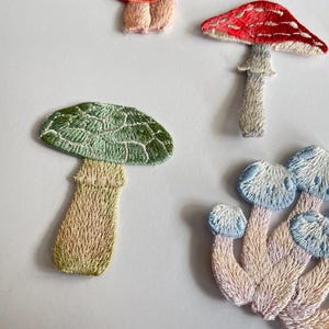 May include: Embroidered mushroom patches in various colors. The patches feature detailed stitching, with a red and white mushroom, a green mushroom, and a cluster of blue and white mushrooms. These patches are ideal for adding a whimsical touch to clothing or accessories.