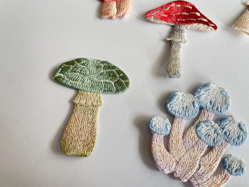 May include: Embroidered mushroom patches in various colors. The patches feature detailed stitching, with a red and white mushroom, a green mushroom, and a cluster of blue and white mushrooms. These patches are ideal for adding a whimsical touch to clothing or accessories.