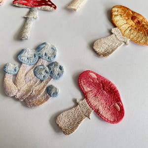May include: Embroidered mushroom patches in various colors, including red, orange, blue, and green. The patches have detailed stitching and are arranged on a white surface. These patches are ideal for adding a whimsical touch to clothing or accessories.