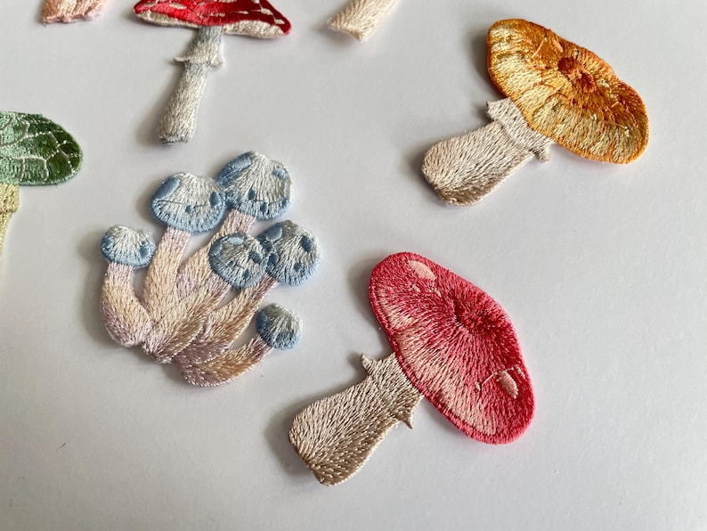 May include: Embroidered mushroom patches in various colors, including red, orange, blue, and green. The patches have detailed stitching and are arranged on a white surface. These patches are ideal for adding a whimsical touch to clothing or accessories.