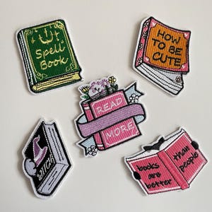 Könnte beinhalten: Gestickte Aufnäher mit Buchmotiven. Ein Aufnäher trägt die Aufschrift "Spell Book" in Grün und Gold. Ein anderer sagt "How to be Cute" in Orange. Andere Aufnäher sagen "Read More" und "books are better than people".