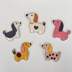 Könnte beinhalten: Fünf gestickte Dackel-Patches in verschiedenen Farben. Einer ist schwarz-weiß, ein anderer rosa, und die anderen sind gelb, hellbraun und weiß. Jeder Patch zeigt ein niedliches Hunde-Design mit Herz- oder Tupfenmuster.