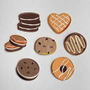 May include: A selection of embroidered patches depicting different cookie designs. The patches include a stack of chocolate sandwich biscuits, heart-shaped biscuits with white icing, and round biscuits with chocolate and vanilla frosting. The patches are in brown, orange, and white.