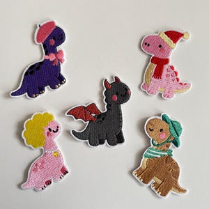 Może przedstawiać: Pięć haftowanych naszywek z dinozaurami w różnych kolorach. Fioletowy dinozaur nosi różowy beret i kokardę. Różowy dinozaur ma czapkę i szalik Świętego Mikołaja. Inne dinozaury są różowe z żółtymi włosami, czarne z czerwonymi skrzydłami i brązowe z zielonym kapeluszem i szalikiem.