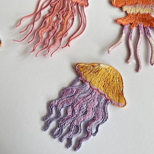May include: Embroidered jellyfish patches in shades of yellow, orange, pink, and purple. The patches feature detailed stitching, with flowing tentacles and a rounded bell shape. These patches are ideal for adding a unique touch to clothing or accessories.
